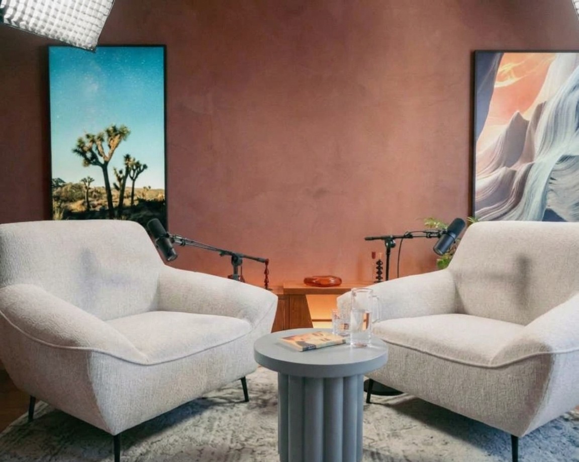 An interview set, empty: two cream armchairs facing each other, microphones on booms, a small round table, and framed desert photographs on a terracotta wall